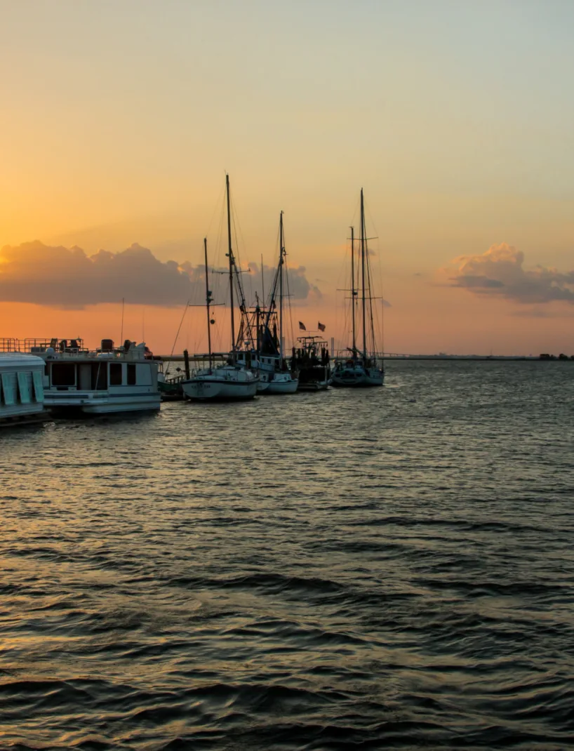 Sunset at a marina with silhouettes of various boats against an orange sky, reflecting on the water.