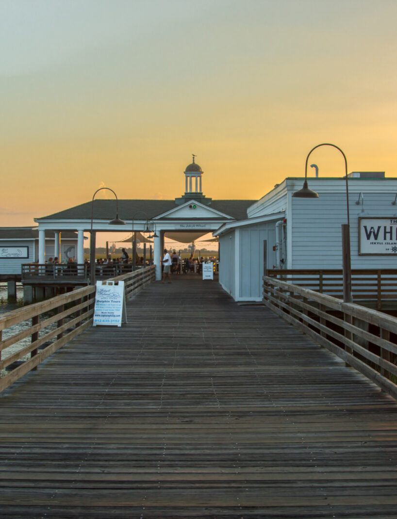 Wooden pier leading to 'The Wharf' restaurant at Jekyll Harbor, with outdoor seating and sunset sky reflecting on the water.