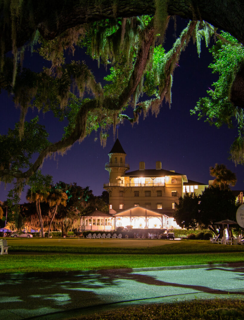 Nighttime shot of Jekyll Island Club Hotel with Spanish moss hanging from oak trees in the foreground. The historic hotel is lit up, with a clear sky above and a 'Do Not Enter' sign to the side.
