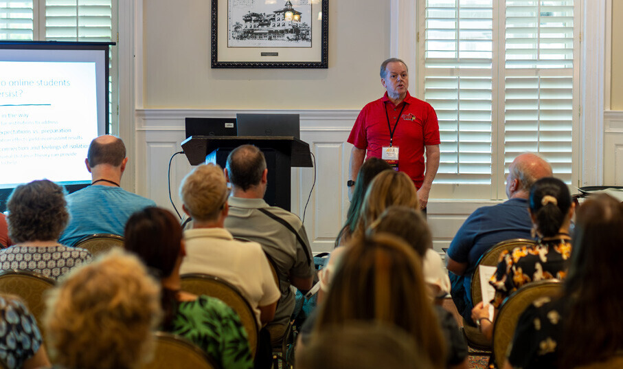 A presenter at dla in a red shirt and badge stands in front of a room, giving a lecture next to a presentation screen. The audience, seated in chairs, is facing the presenter, attentively listening to the speech. The room has bright lighting and a traditional decor.