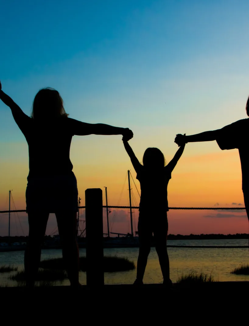 A silhouette of three people holding hands with arms raised, standing in front of a calm waterfront at sunset, with sailboats and a serene sky in the background.