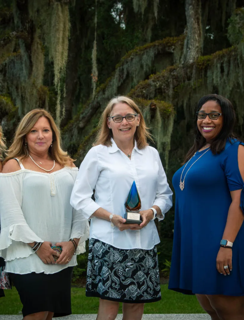 A group of five women stands together outdoors, smiling at the camera. The woman in the center holds an award, with moss-covered trees in the background.