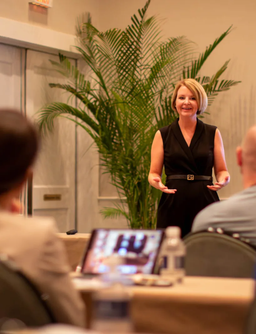 A woman in professional attire stands in front of a room speaking to an audience during the DLA conference, with a palm plant in the background and attendees seated at tables.