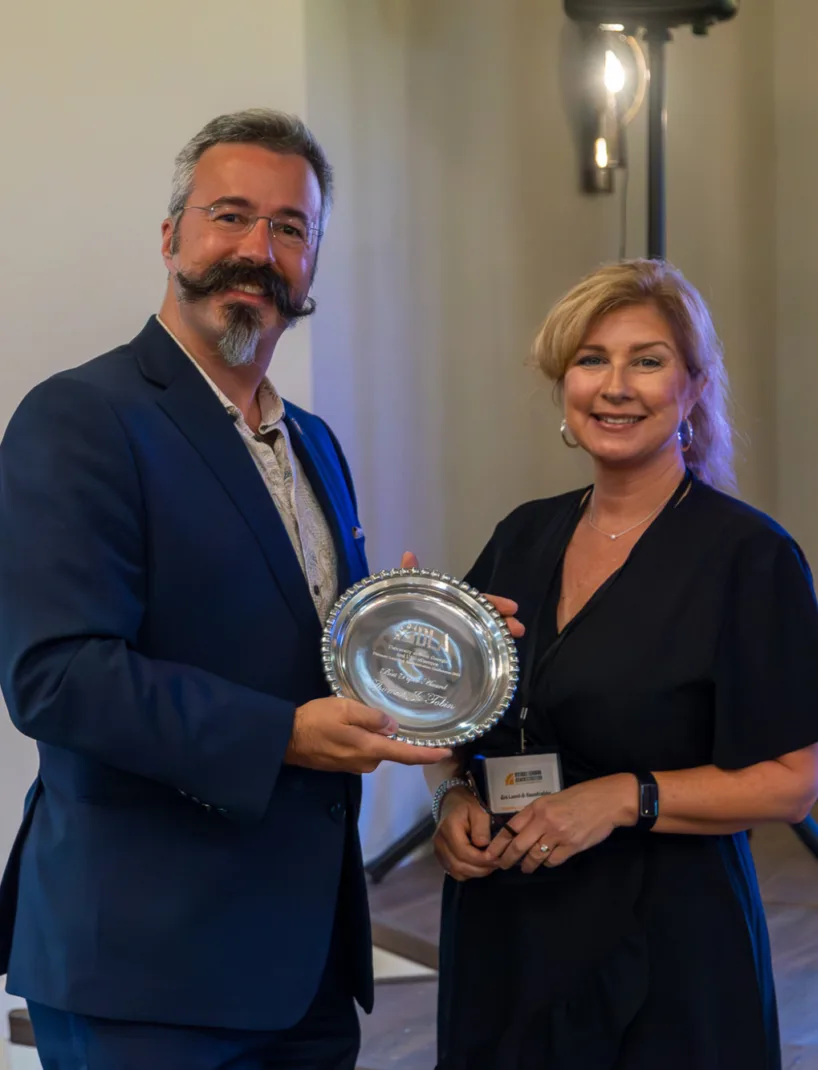 A man in a blue suit holds a silver award plate while standing next to a woman in a dark dress, both smiling at the camera during an award ceremony.