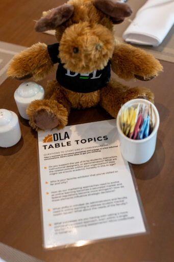A brown stuffed moose in a DLA shirt sits on a table beside a “DLA Table Topics” card, saltshakers, and colorful sugar packets.