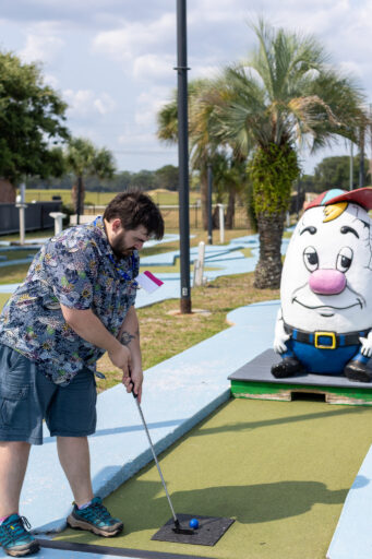 A man lines up a shot with a golf club in a golf course filled with palm trees and fun statues.