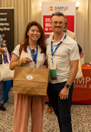A woman and man pose with a Jekyll Island Club Resort bag as people smile in the background.