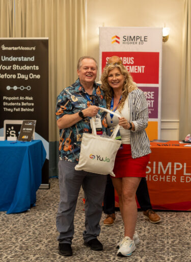 A woman and man pose with a YuJa tote bag as people smile in the background.