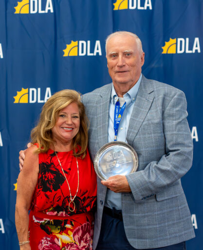 In front of a DLA backdrop, a woman stands next to a man holding a DLA award.