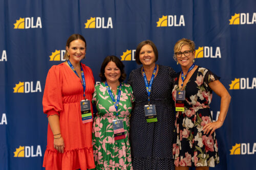 Four women wearing DLA Staff badges are posing in front of a DLA backdrop.