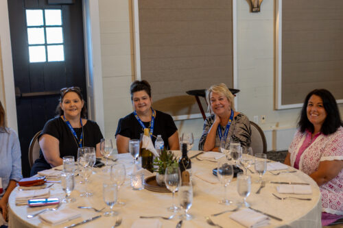 Four women wearing DLA lanyards smile at the camera, while sitting at a round table set with glasses and cutlery.