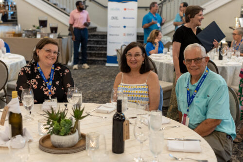 Two women and one man are sitting at a round table facing the camera, while people converse in the background.