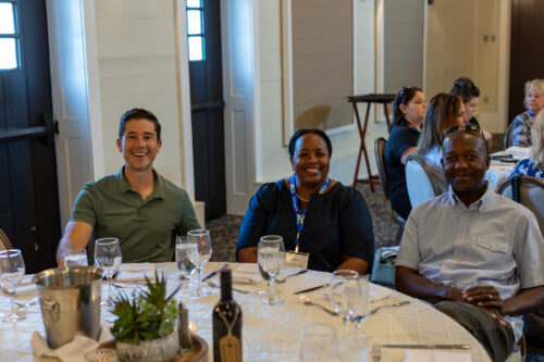 Two men and a woman are smiling at the camera, sitting at a round dinner table. Another round table filled with people is in the background.
