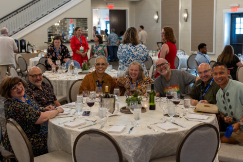 A group of people sitting at a round dinner table smile for the camera. More people are either moving or conversing in the background.
