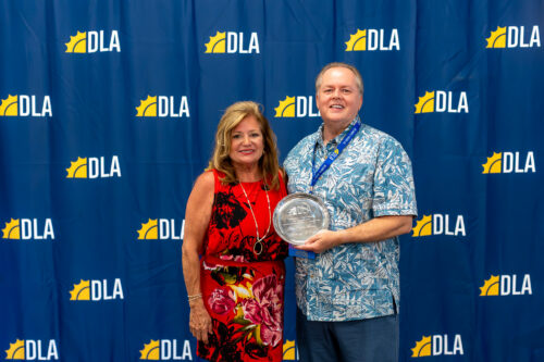 In front of a DLA backdrop, a woman stands next to a man holding a DLA award.