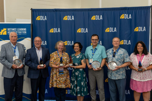 A group of seven people stand in front of a DLA backdrop. Each of them holding an award.