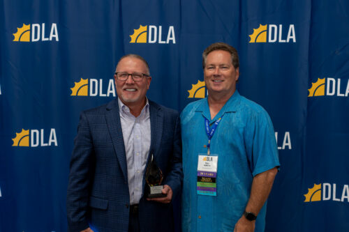 A man holding an award stands next to another man wearing a DLA lanyard in front of a DLA backdrop.