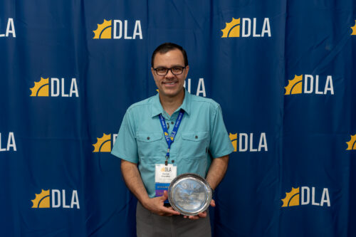 A man stands in front of a DLA backdrop holding an award.