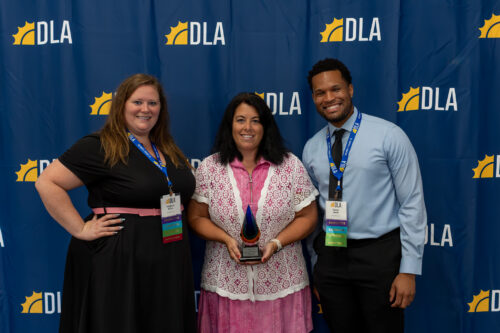 In front of a DLA backdrop, a man and woman stand on opposite sides of a woman holding an award.