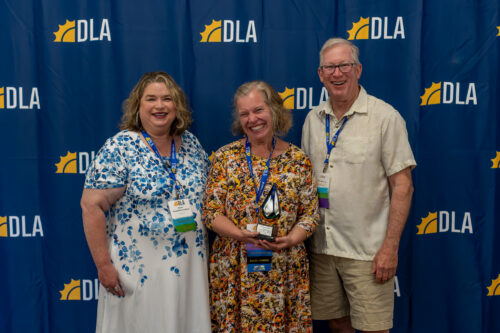 In front of a DLA backdrop, a man and woman stand on opposite sides of a woman holding an award.