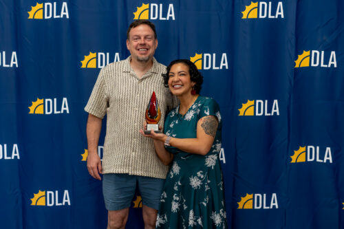 A woman holding an award stands next to a man in front of a DLA backdrop.