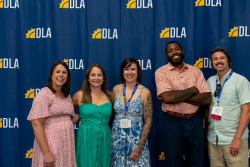 Three women and two men stand, smiling in front of a DLA backdrop.