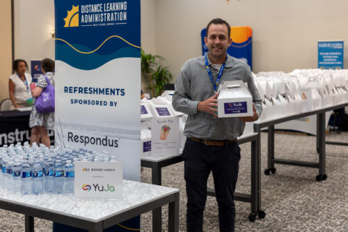 A man holds a lunchbox, posing in front of tables filled with lunchboxes behind a DLA Refreshments sign.