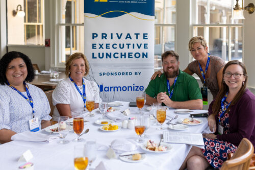 A group of people sitting at a table, pose for the camera in front of a Private Executive Luncheon banner.