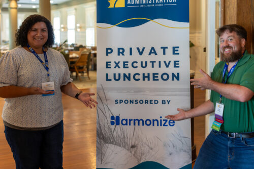 Two people gesture towards a DLA Private Executive Luncheon banner outside a dining room.