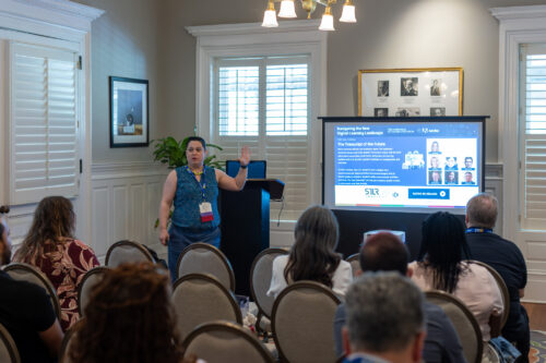 A woman, standing in front of a podium next to a slideshow, raises her hand in front of a seated audience.