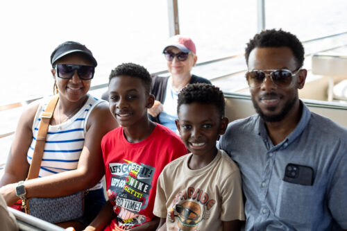 Parents and their two sons sit on a boat bench smiling at the camera.