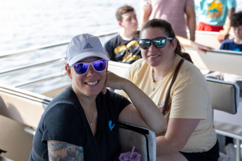 Two women sit on a boat bench smiling at the camera.