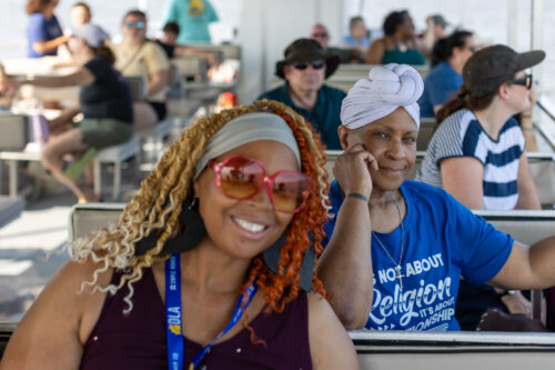 The camera focuses on a woman smiling at the camera with other people smiling or looking over the boats edge in the blurred background.