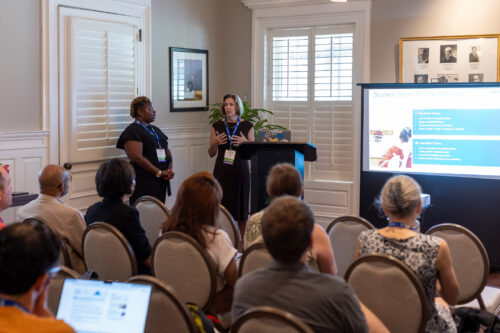 A woman standing beside a podium and slideshow gives a presentation to a seated audience.