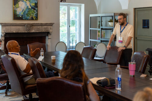 A group meets around a conference table in an elegant room; a man stands looking at the seated speaker as other participants sit listen attentively.