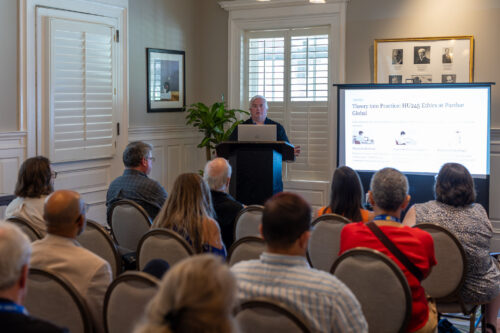 A man, standing behind a podium next to a slideshow, speaks to a seated audience.