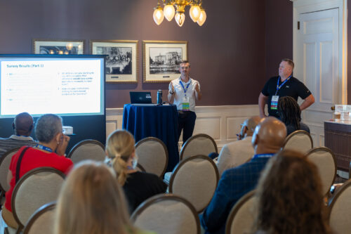 A man, standing behind a table next to a slideshow, speaks to a seated audience.