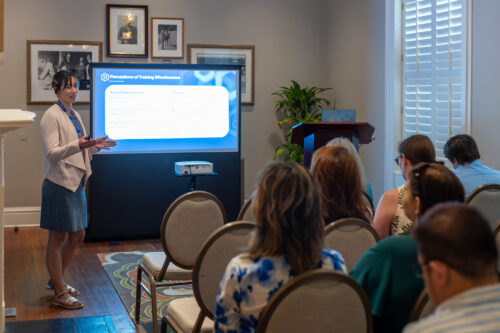 A woman, standing next to a slideshow, speaks to a seated audience.