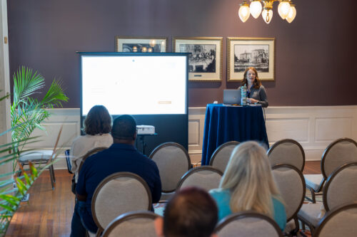 A woman, standing behind a table next to a slideshow, speaks to a seated audience.