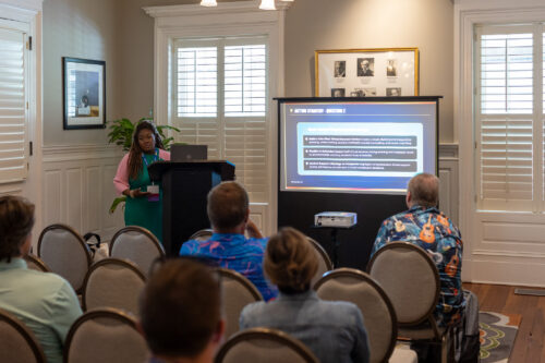 A woman, standing behind a podium next to a slideshow, speaks to a seated audience.