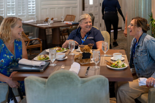 Three women sit laughing at a dinner table filled with salads and glasses of water.