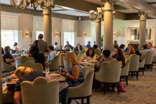 Elegant dining room filled with guests enjoying meals, framed by ornate columns, chandeliers, and bright windows.