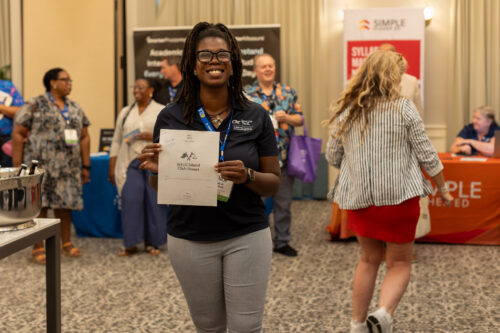 Standing in front of booths, a woman wearing a DLA lanyard and holding a Jekyll Island and Club Resort flyer smiles at the camera.