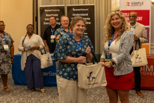 Two women pose with a Honorlock tote bag as people smile in the background.