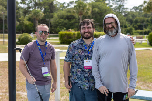 Three men wearing DLA lanyards in a greenery setting hold golf clubs while smiling for the camera.