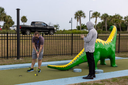 A man stands beside a colorful dinosaur watching another man try to hit a golf ball into a hole.