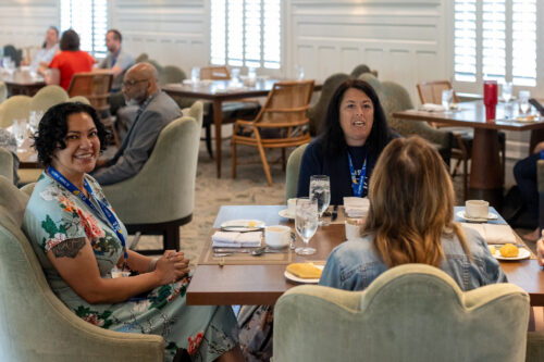 A group of women sitting at a square dinner table. More people are conversing in the background.