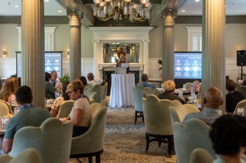 A woman, standing behind a podium in between two slideshow displays, reads off a sheet of paper to her seated audience in a dining room.