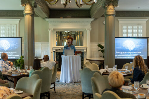 A woman, standing behind a podium in between two slideshow displays, speaks to a seated audience.