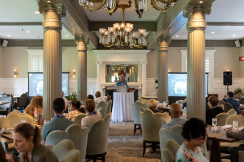 A woman, standing behind a podium in between two slideshow displays, speaks to a seated audience in a dining room.